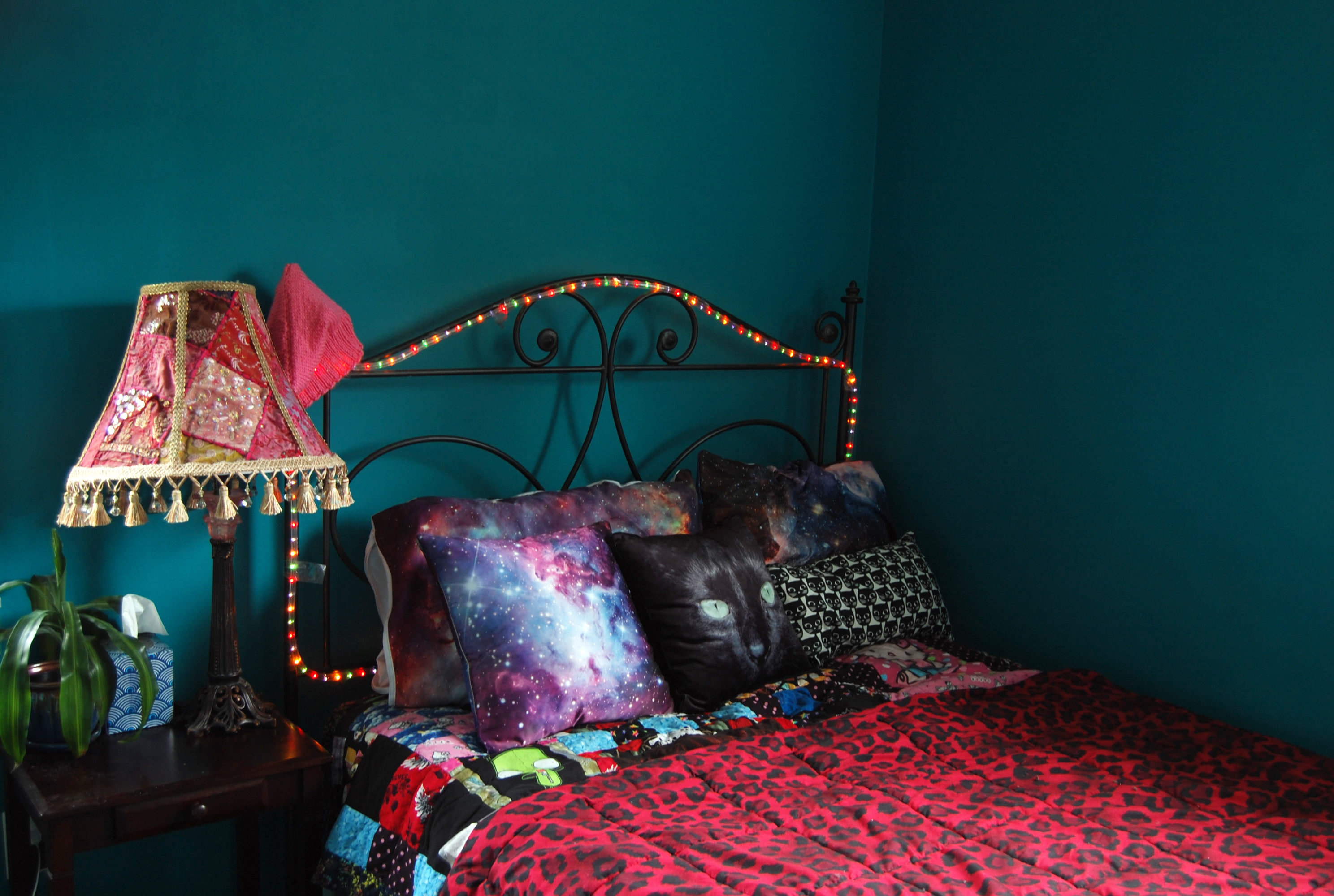 A quirky made bed with lots of pillows next to a colorful lamp in a teal bedroom