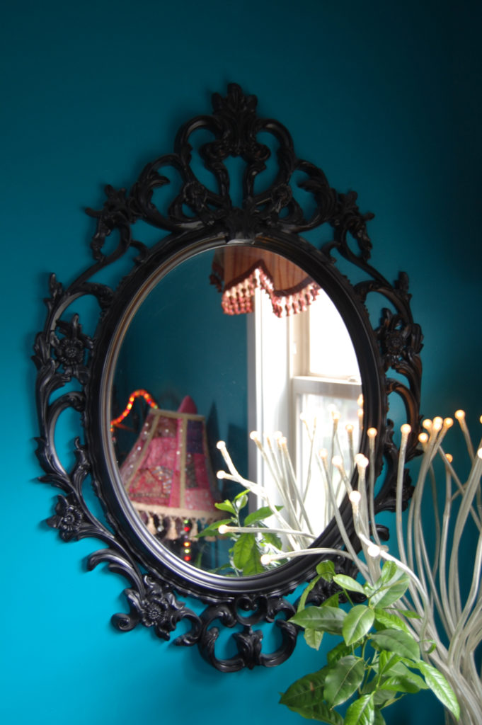 A black baroque-framed mirror with a plant and a lamp that looks like brain neurons in a teal bedroom