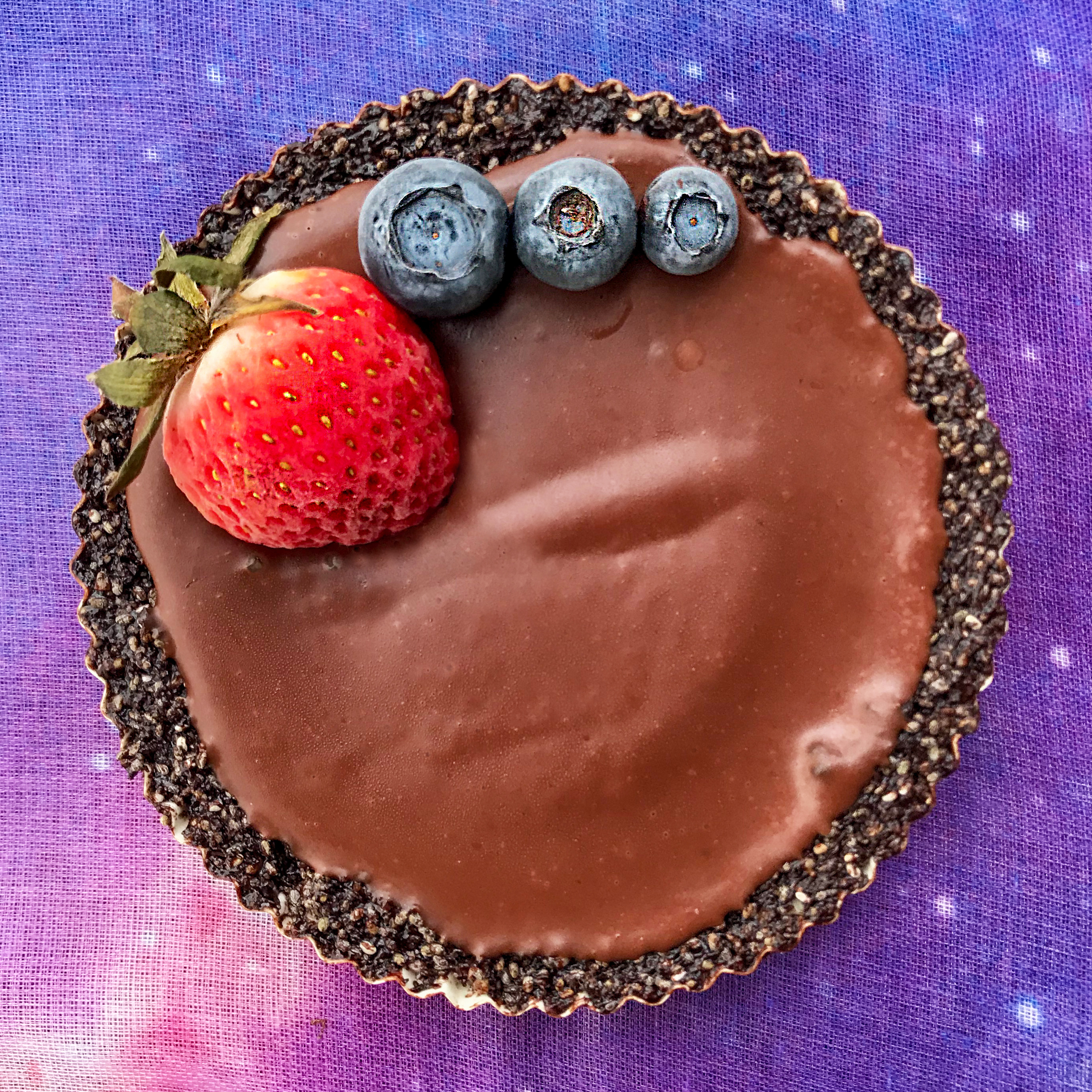 A round 4-inch vegan chocolate tarte with a cashew-chia seed crust, plus a strawberry and 3 blueberries as a garnish.