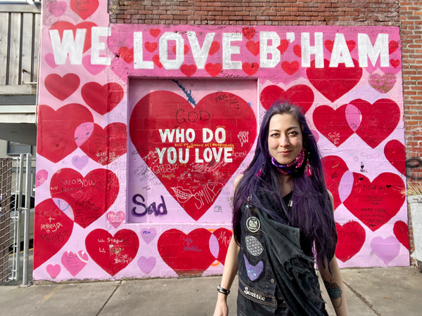 Ronnie stands in front of a pink mural with red hearts that reads, "We Love B'Ham."