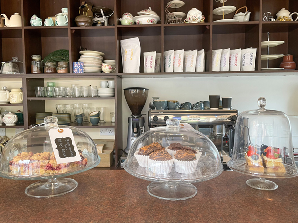 An assortment of cakes display under bell jars on the front counter, in front of a shelf holding teacups with floral patters.