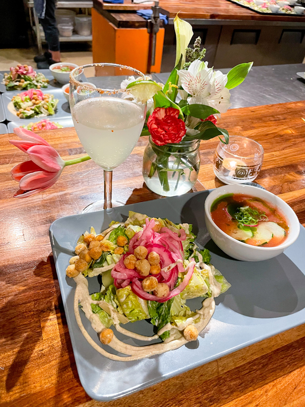 A romaine Caesar salad with chickpea croutons and pickled red onion is plated next to a small bowl of red and green cold gazpacho soup. Behind the plate is a lemon-lime elixir next to a small glass of fresh-cut flowers and a lit tea candle.