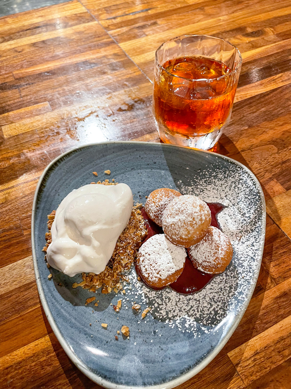 Four bite-sized beignets dusted with powdered sugar sit on top of a pool of elderberry puree. Next to the beignets is a scoop of coconut-based ice cream on top of crushed granola. A small glass of iced tea is behind the dish.