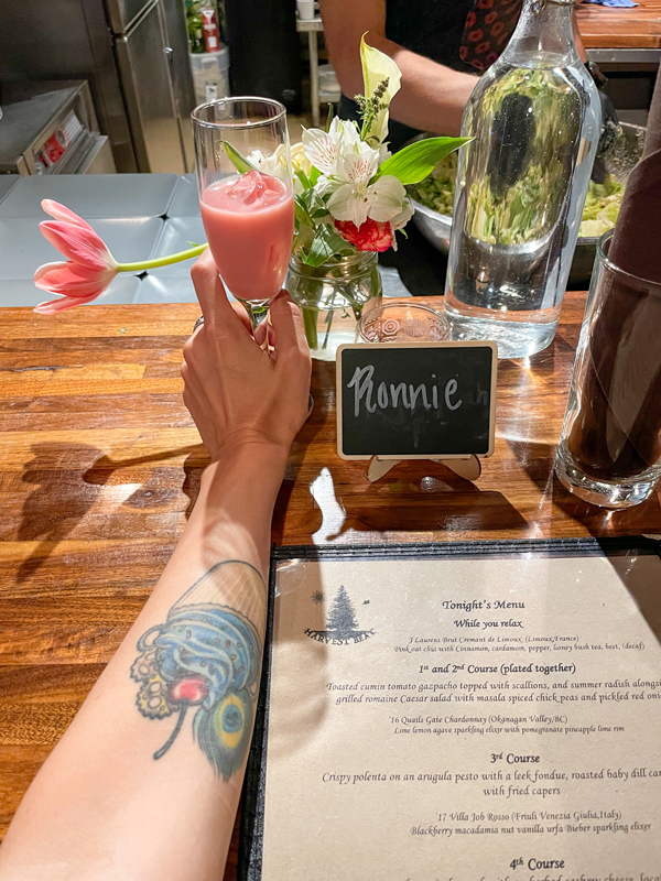 Ronnie's left hand with a cupcake tattoo on her forearm reaches for the pink oat chai drink. On the right is Ronnie's name card with the evening multi-course menu in front of it.