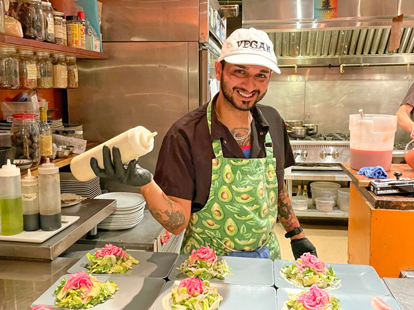 With the Caesar dressing bottle in hand, Karim smiles at the viewer as he prepares numerous Caesar salads.